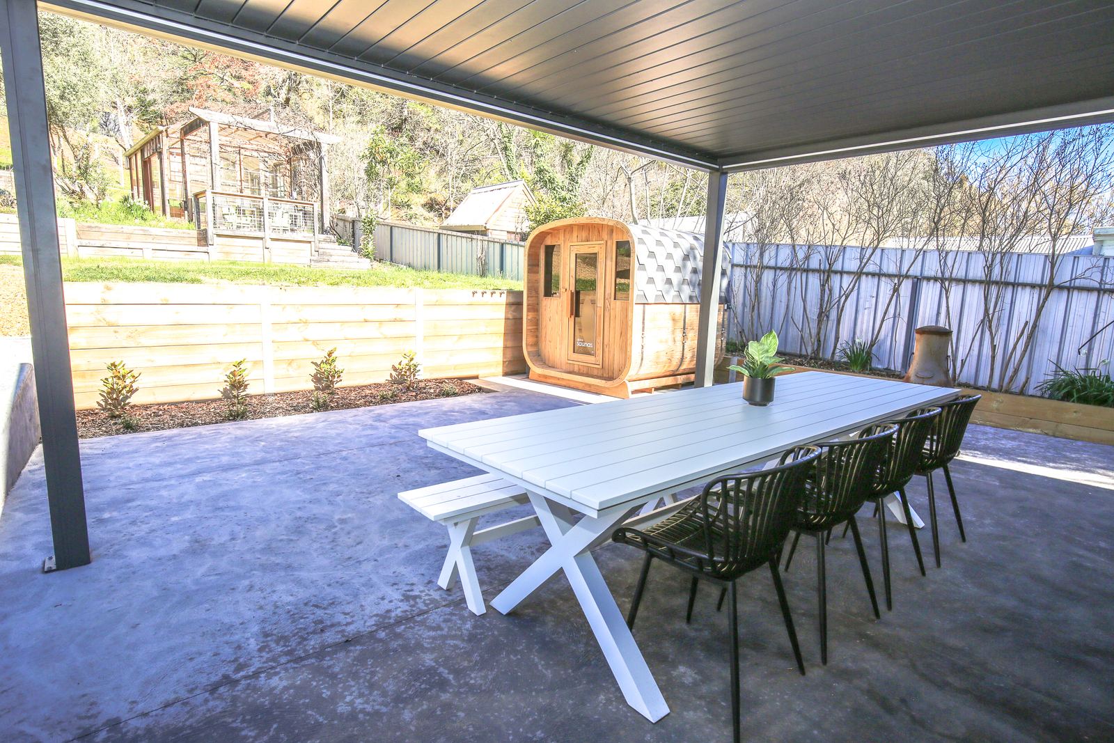 The covered alfresco area with the long white outdoor table and chairs, the sauna in view across the terrace