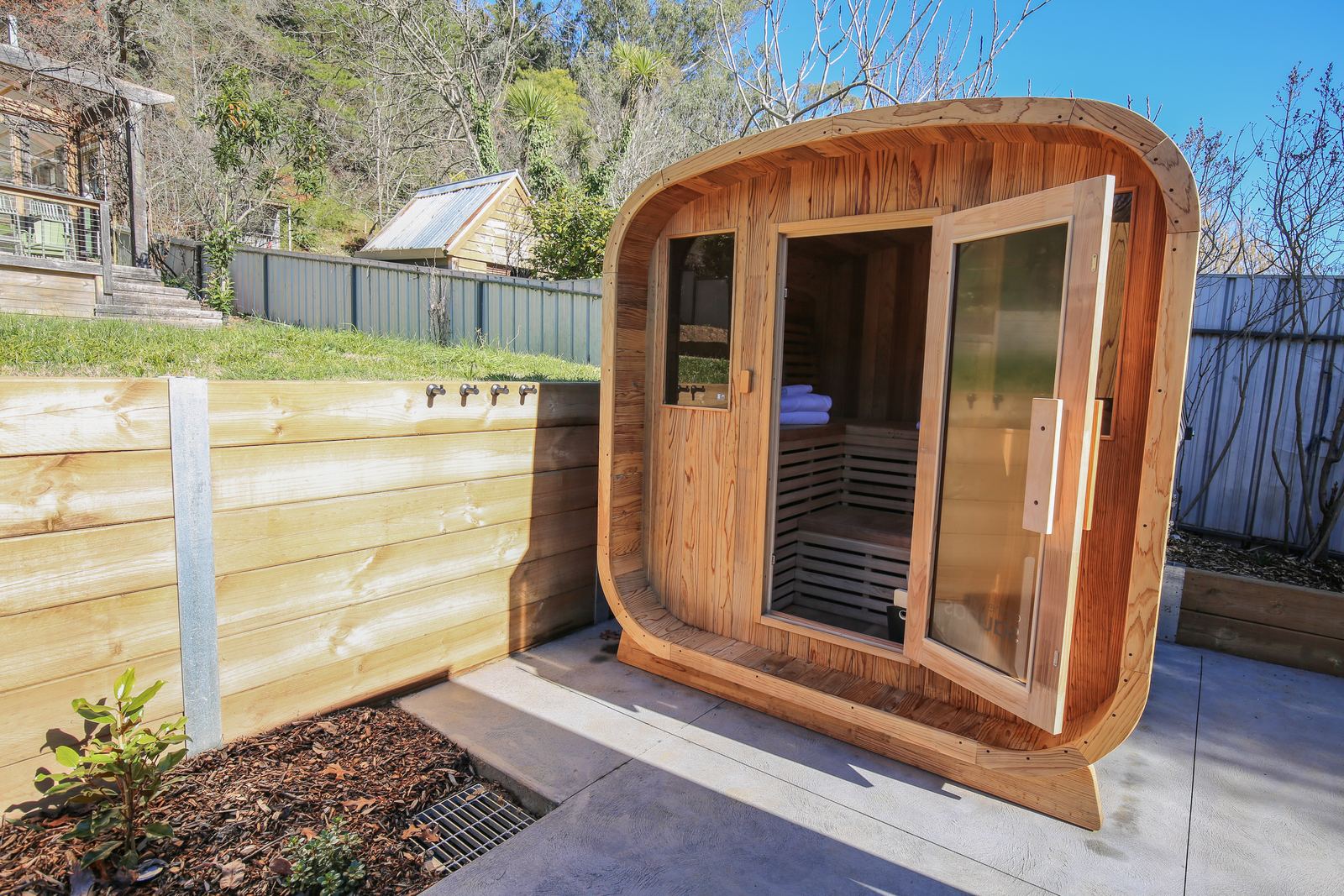 The private outdoor sauna at Stoke and Steam, a rounded timber cabin on the paved terrace, its glass door open with folded towels on the bench inside