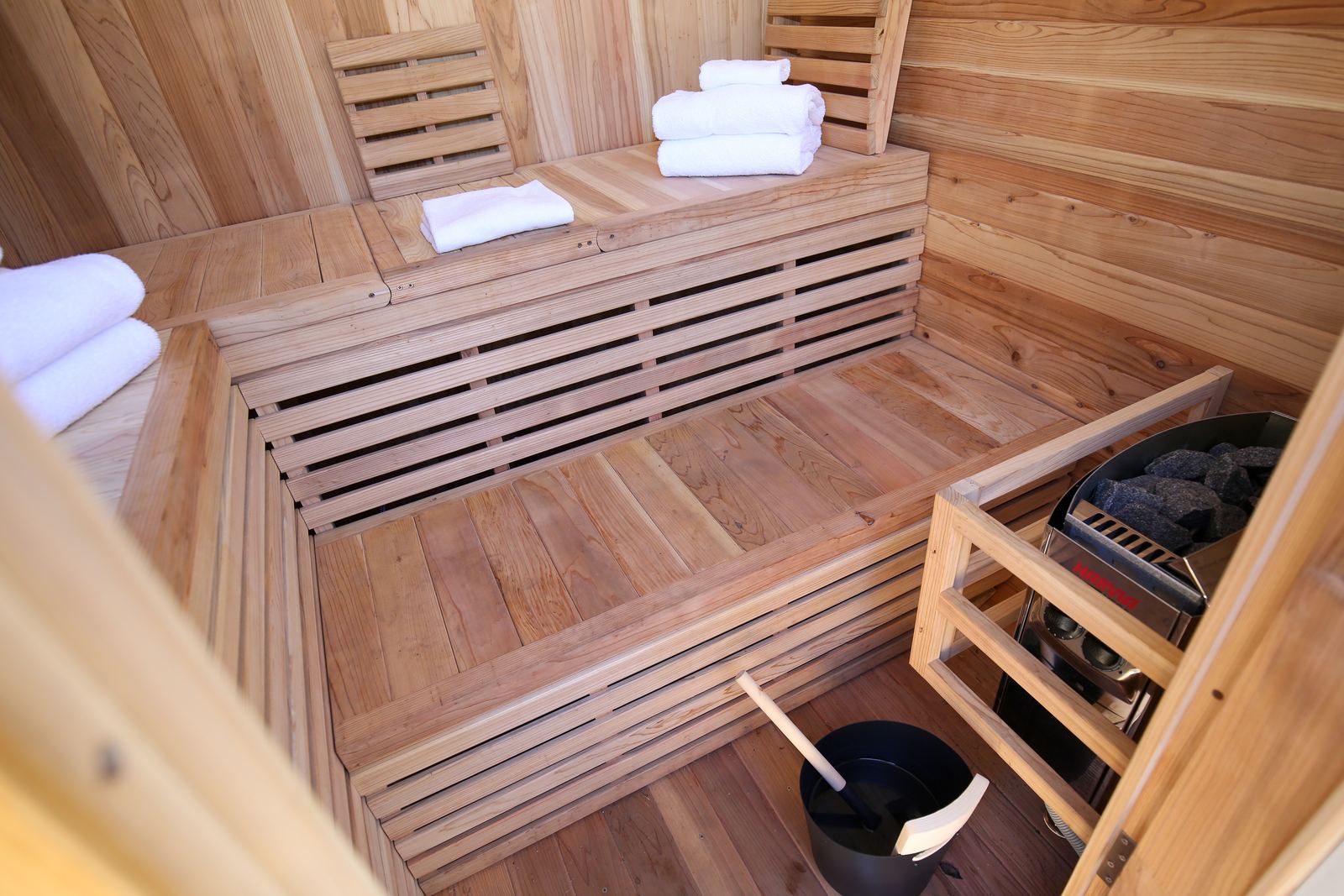 Inside the sauna: two tiers of timber benches with folded towels, the electric heater with its stones, and a bucket and ladle on the floor